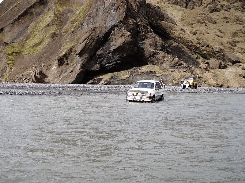 White SUV fording a wide gray river with mountains behind