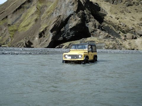 1996 Ford Bronco - A yellow Land Rover Defender navigates a shallow river, with , rocky terrain rising behind it.