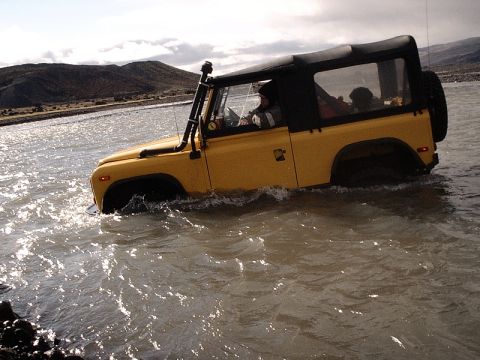 1996 Ford Bronco - Off-road vehicle navigates a shallow river under a cloudy sky.