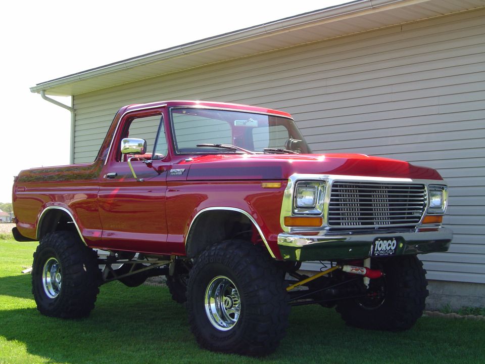 Lifted red 1978 Ford Bronco on chrome wheels and big tires, 3/4 front view on grass.