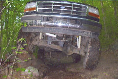 1995 Ford Bronco - A black Ford truck navigates a rocky, wooded trail, showing its front grille and suspension.