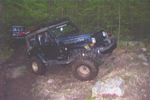 1995 Ford Bronco - Two off-road vehicles navigate a rocky, wooded trail, with the lead Jeep Wrangler climbing over obstacles.