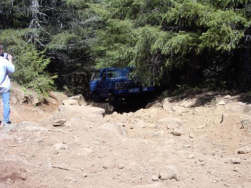 Blue lifted Ford truck descending a rocky forest trail as a bystander photographs