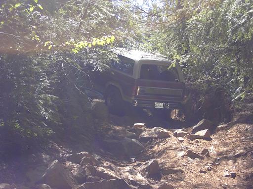 Two-tone full-size SUV descending a rocky forest trail between dense evergreens, rear quarter view