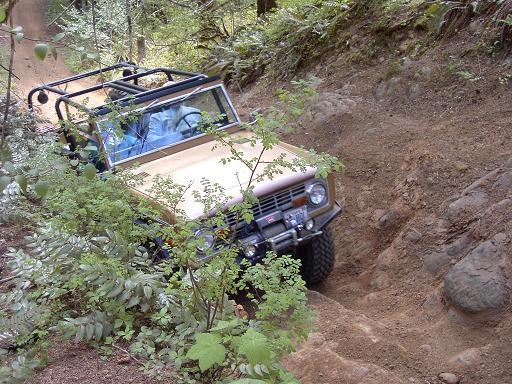 Tan open-top classic-style Ford Bronco with roll cage and winch descending a rocky forest trail