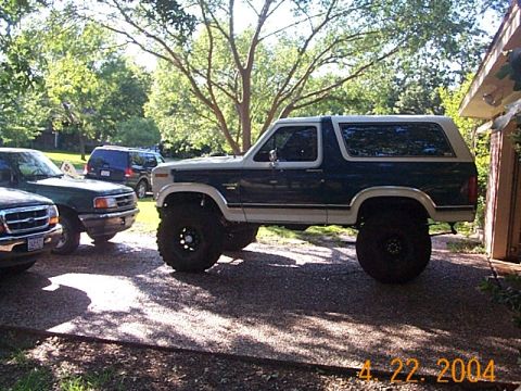 1986 Ford Bronco - This lifted two-tone SUV, likely a 1980s Ford Bronco, is parked outdoors next to other vehicles.
