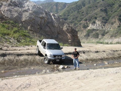 1985 Ford Bronco - A man stands beside a lifted pickup truck navigating a rocky, dry riverbed.