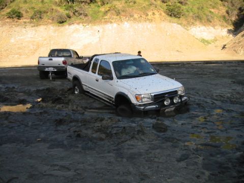 1985 Ford Bronco - Two pickup trucks are stuck in thick mud on a dirt road with hills in the background.