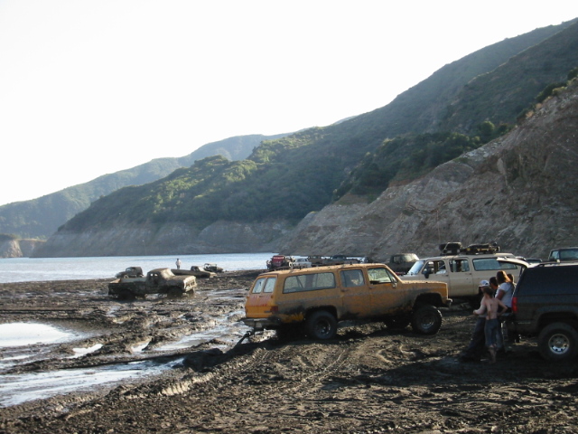 Group of off-road SUVs parked on a muddy lakeshore beneath green coastal hills