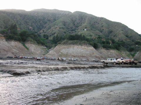 1985 Ford Bronco - A collection of trucks and SUVs are gathered in a muddy, outdoor area with hills visible in the distance.