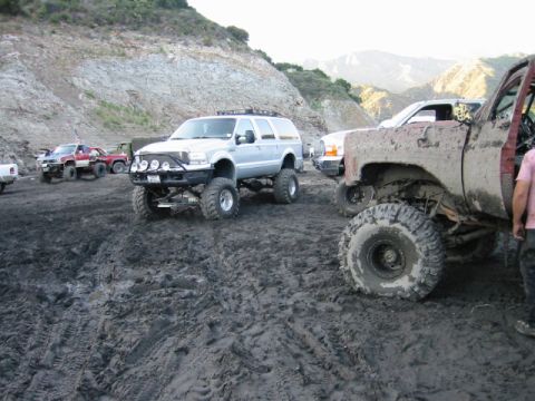 1985 Ford Bronco - Multiple modified off-road vehicles are stuck in a muddy terrain with hills in the background.