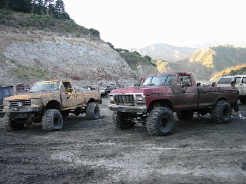 1985 Ford Bronco - Two heavily modified, mud-splattered pickup trucks are parked on a dirt lot with a rocky hillside in the background.
