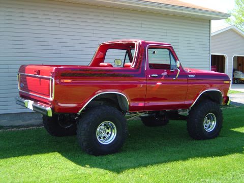 1978 Ford Bronco - red lifted classic Ford Bronco with chrome wheels and mud tires, parked on grass beside a house and garage.