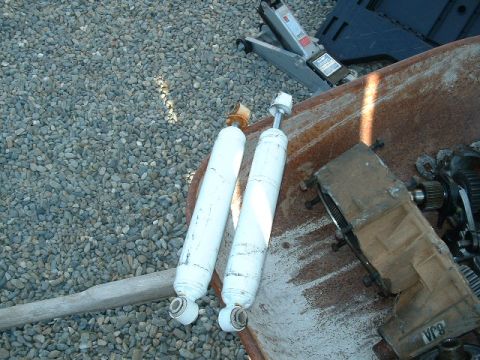 1991 Ford Bronco - Two white shock absorbers and transmission components are laid out on a rusty surface with gravel and a car jack in the background.