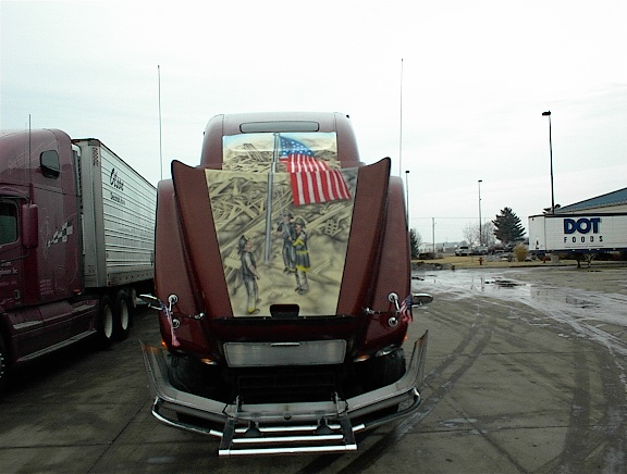 Semi-truck hood painted with 9/11 theme, showing firefighters and American flag. Another semi-truck is parked to the left