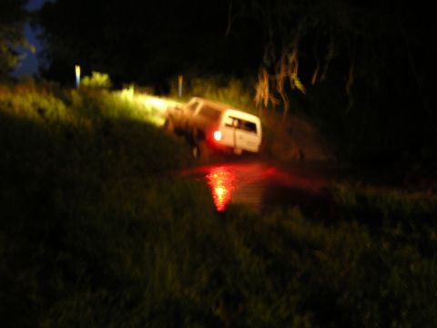 1984 Chevrolet Blazer - A white SUV is partially submerged in water on a grassy slope at night, its taillights casting a red reflection.