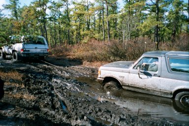 1979 Ford Bronco II - Two off-road vehicles are stuck in a muddy trail surrounded by trees.