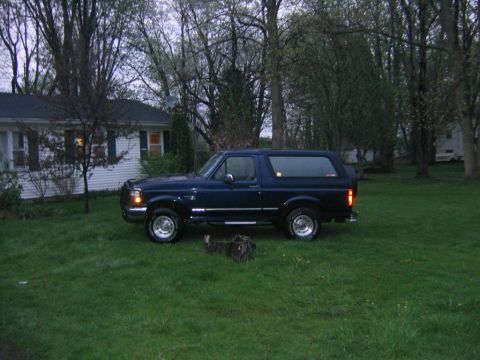 1994 Ford Bronco - A dark blue SUV is parked on a lawn in front of a house, with trees surrounding the property.