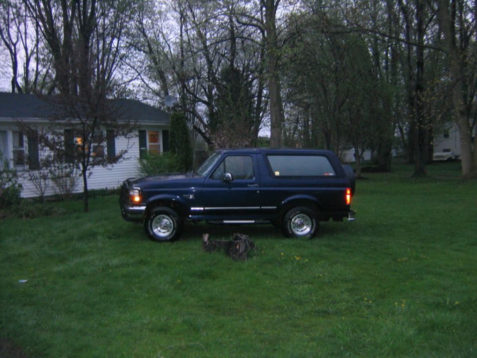 Dark blue Ford Bronco parked on a lawn at dusk with lights on, driver-side profile view