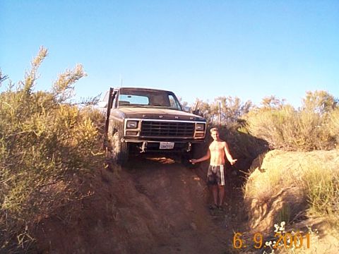 1981 Ford Bronco - Front view of a dark, mud-splattered square-body Ford Bronco with FORD lettering on the hood grille panel, nosed down a rutted dirt trail c…