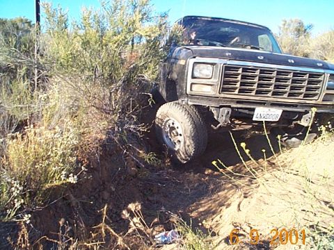1981 Ford Bronco - Low front three-quarter shot of a black square-body Ford Bronco flexing on a rutted dirt trail, its left front tire lifted over an eroded b…