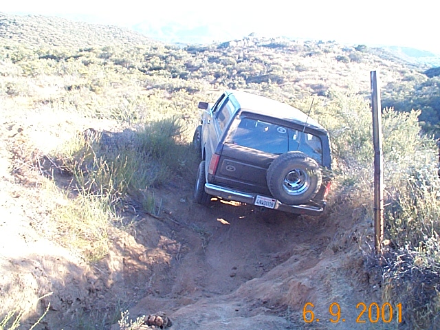Black Ford Bronco with white roof descending a steep rutted dirt hill, rear three-quarter view