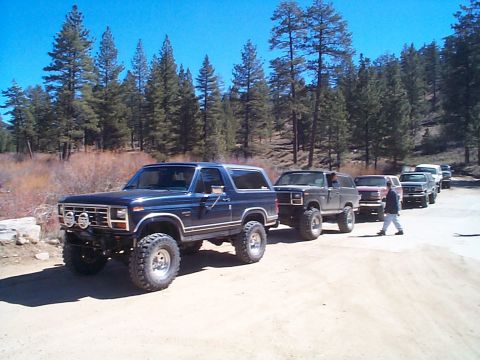 1981 Ford Bronco - A lifted dark blue square-body Ford Bronco with a brush guard, KC-style front lights, chrome wheels and tall mud-terrain tires leads a line…