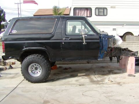 1981 Ford Bronco - Side profile of a black square-body Ford Bronco parked on a driveway mid-teardown, with the hood, front fenders, grille and front wheel str…