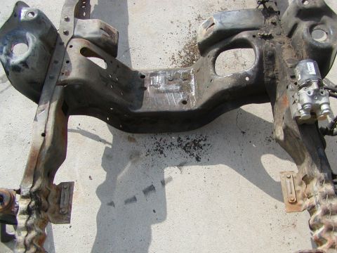 1981 Ford Bronco - A section of a vehicle's frame, showing rust and attached hydraulic parts, rests on a concrete surface.