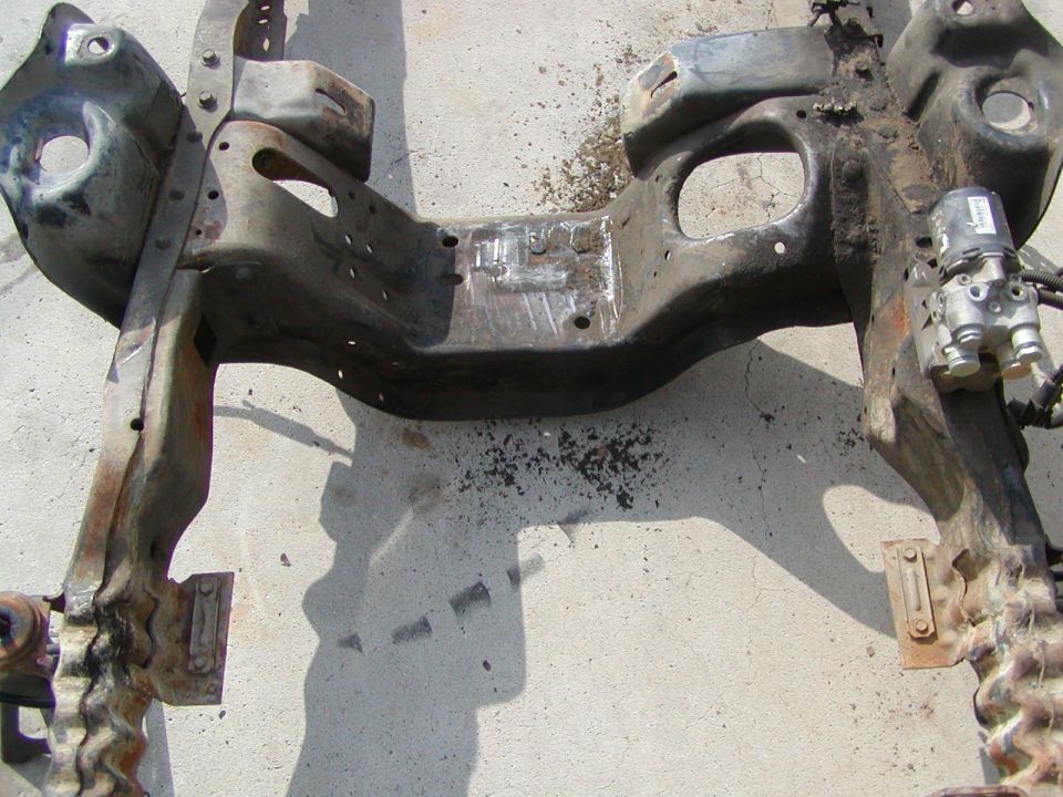 Rusty metal vehicle frame section with hydraulic components attached, lying on concrete