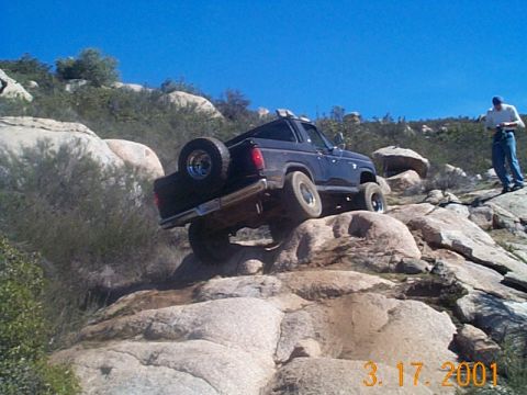 1981 Ford Bronco - Blue-gray full-size Bronco with the top removed climbing a granite ledge with the driver-side rear wheel lifted well off the rock, dated 3-…