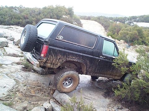 1981 Ford Bronco - Black full-size Bronco with removable hardtop flexing over a granite slab on a rocky trail, mud caked up the rear quarter and rocker. Rear-…