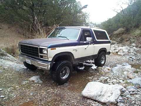 1981 Ford Bronco - Blue-and-cream two-tone full-size Bronco with a white hardtop sitting in a rocky creek bed, shot from the driver-side front three-quarter.…