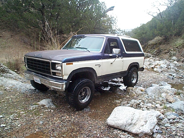 Blue and cream Ford Bronco parked in a rocky creek bed, front 3/4 view, lifted on mud tires