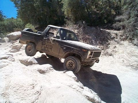 1981 Ford Bronco - Dust-coated black full-size Bronco with the top removed and a white roll bar, dropping down a granite ledge on a pine-lined trail with the…