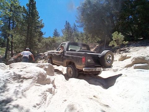 1981 Ford Bronco - Black topless full-size Bronco with a roll bar and rear-mounted spare crawling over pale granite slabs on a pine-shaded mountain trail, dus…
