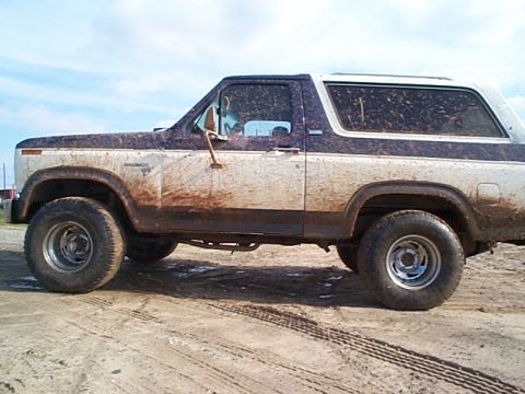 1981 Ford Bronco - Driver-side profile of a two-tone dark-over-white square-body Ford Bronco caked in dried mud from rocker to roofline, parked on a rutted di…