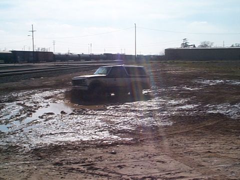 1981 Ford Bronco - Distant three-quarter front view of a light-colored square-body full-size Bronco-style SUV sitting in a muddy, rutted open lot full of wate…