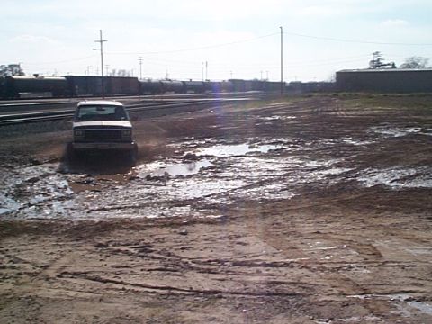 1981 Ford Bronco - A white SUV navigates a muddy terrain with train cars in the background.