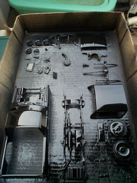 1981 Ford Bronco - Freshly painted gloss-black scale model truck parts laid out to dry in a cardboard box, including the cab and bed, a ladder frame with axle…