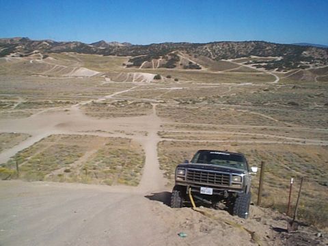 1981 Ford Bronco - Black lifted full-size Bronco with a black grille and light bar climbing a sandy berm at a desert OHV area, yellow tow strap hanging from t…
