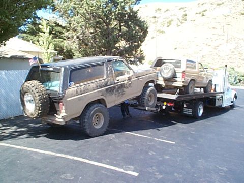 1981 Ford Bronco - Mud-covered black full-size Bronco with an American flag on the rear spare carrier being winched or backed onto a flatbed tow truck in a pa…