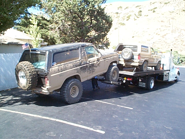 Muddy black Ford Bronco being loaded onto a flatbed tow truck with another Bronco aboard
