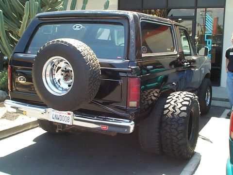 1981 Ford Bronco - Glossy black full-size Bronco parked at an Alpine car-audio storefront, shot from the driver-side rear three-quarter with the swing-out spa…