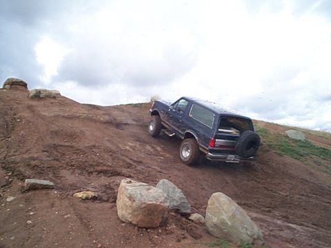 1981 Ford Bronco - Dark blue full-size Bronco with a rear-mounted spare climbing a steep red dirt hill, front wheels throwing mud under an overcast sky. Slott…