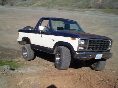 1981 Ford Bronco - Front three-quarter view of a dark blue and white two-tone full-size Ford Bronco with the rear top removed, parked on a dirt pullout beside…