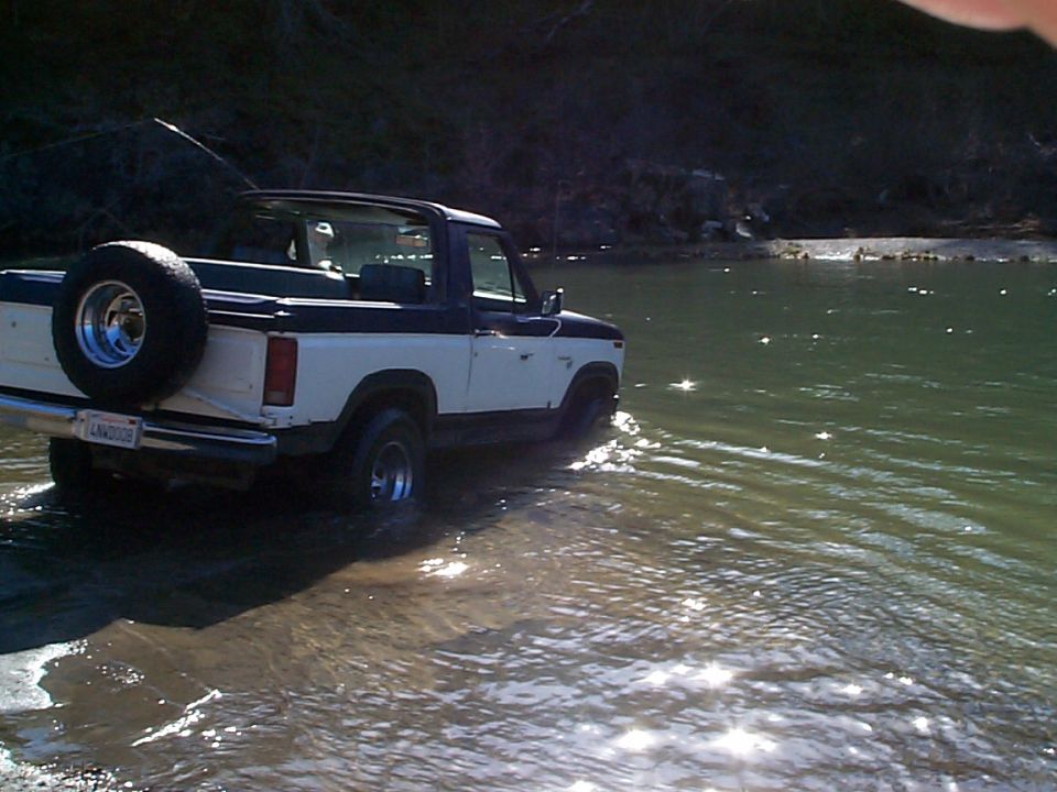 Two-tone white and blue topless Ford Bronco fording a shallow river, rear three-quarter view