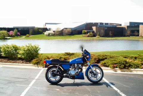 1985 Ford Crown Victoria LTD - A blue Suzuki motorcycle is parked in a lot, with a lake and office buildings visible behind it.