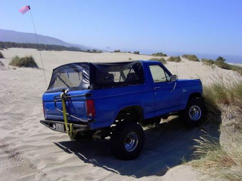 1980 Ford Bronco - Blue 1980 Ford Bronco with soft top on sand dunes, rear-mounted spare tire carrier visible.