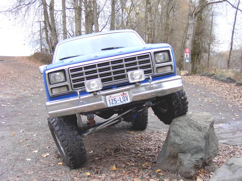 Lifted blue 1980s Ford 4x4 flexing front suspension over a boulder on gravel road